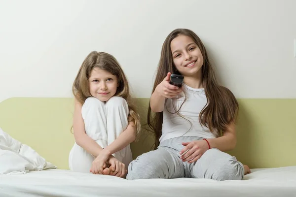 Niños sentados en casa en la cama y viendo la televisión. En las manos sosteniendo control remoto de televisión —  Fotos de Stock
