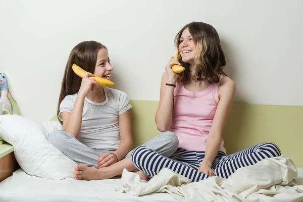 Двое детей-сестер в пижаме играют по утрам в постели. Держите бананы как телефоны, говорящие и смеющиеся . — стоковое фото
