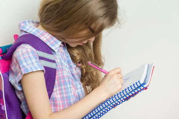 Niña estudiante de escuela primaria con gafas con una mochila escribiendo en su cuaderno . — Foto de Stock