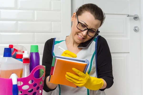 Dienstmeid vrouw is het reinigen. Vrouw in gele rubberen handschoenen, praten over telefoon schrijven in notitieblok. — Stockfoto