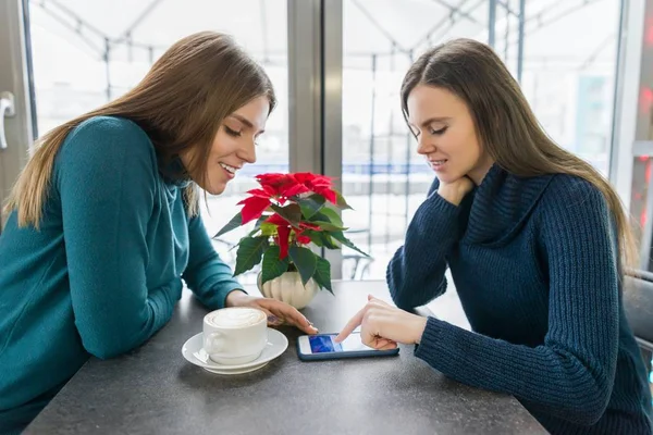 Talking young women, girls sitting in winter cafe smiling and talking using smartphone — Stock Photo, Image