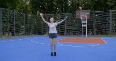 Hareketli, gülümseyen, mor saçlı, kolları havada, sokak basketbol sahasında zıplayan bir genç kız.