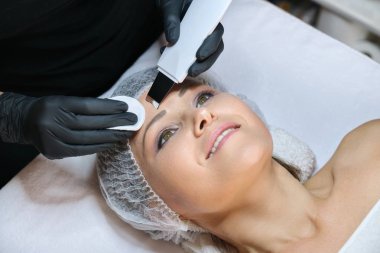 Kozmetoloji, olgun bir kadın ultrasonik bir aletle yüz temizliği yaptırıyor.
