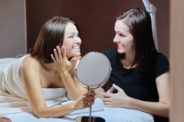 Falando e sorrindo mulheres esteticista e maduro paciente feminino com espelho no salão de beleza — Fotografia de Stock