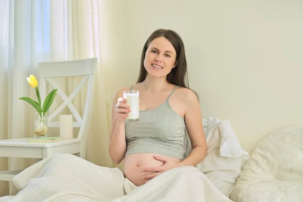 Mladá usměvavá těhotná žena se sklenicí mléka sedí doma v posteli — Stock fotografie