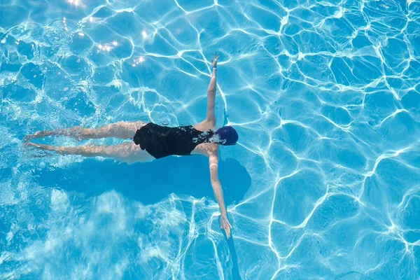 Νεαρή γυναίκα σε καπέλο σπορ μαγιό που κολυμπά κάτω από το νερό σε μπλε εξωτερική πισίνα — Φωτογραφία Αρχείου