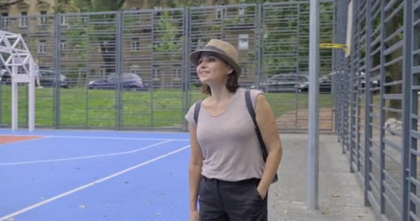 Dojrzała kobieta z plecakiem w kapeluszu przyszła do pustego boiska do koszykówki sportowej — Wideo stockowe