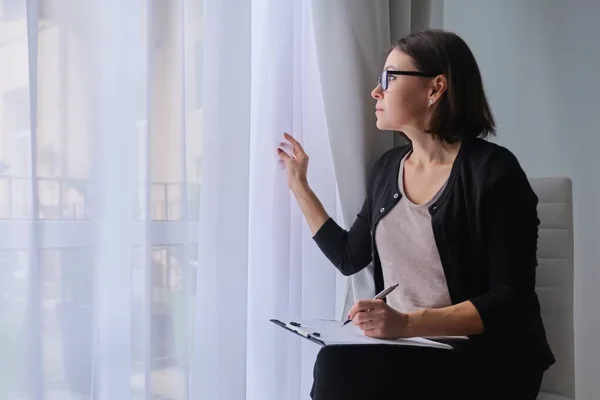 Mujer madura trabajadora social, psicóloga sentada cerca de la ventana con portapapeles — Foto de Stock