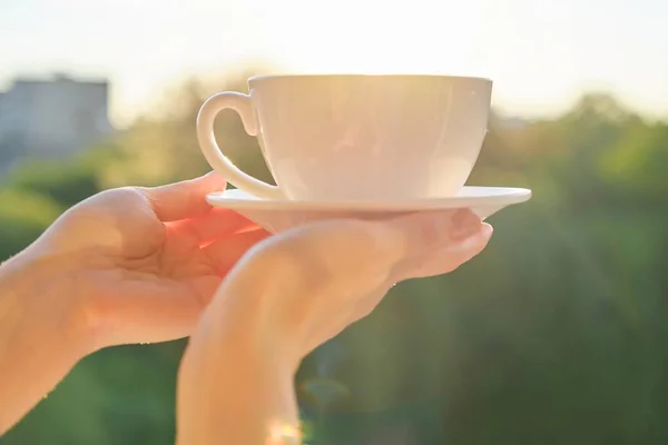 女性の手でソーサーと白いセラミックカップのセット,背景の夕日 — ストック写真