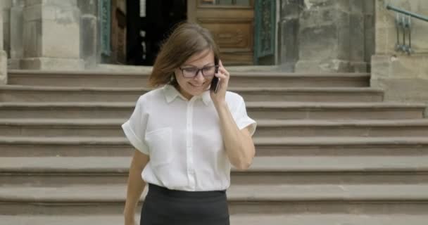 Ältere lächelnde positive Frau mit Brille, helle Bluse, die auf der Treppe läuft und telefoniert — Stockvideo