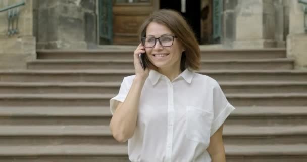 Madura sonriente mujer positiva en gafas blusa ligera caminando por las escaleras hablando por teléfono — Vídeos de Stock