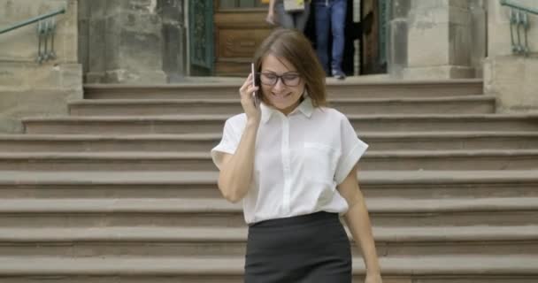 Madura sonriente mujer positiva en gafas blusa ligera caminando por las escaleras hablando por teléfono — Vídeos de Stock