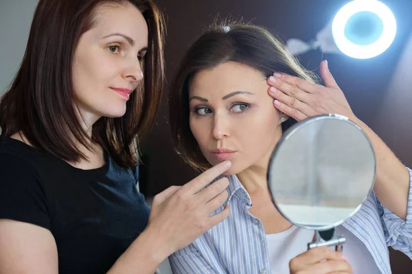 Paciente feminina de meia-idade olhando no espelho e esteticista feminina mostrando no rosto — Fotografia de Stock