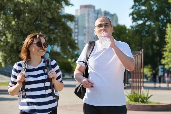 Maduro sonriente hombre y mujer caminando en el parque de la ciudad hablando de agua potable de la botella —  Fotos de Stock