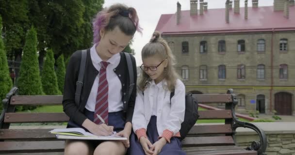 Две школьницы, сидящие на скамейке, учащиеся начальной и средней школы . — стоковое видео