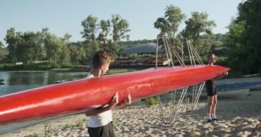 Genç bir delikanlı spor kanosuyla, gençler kollarında tekneyle nehre doğru yürüyorlar.