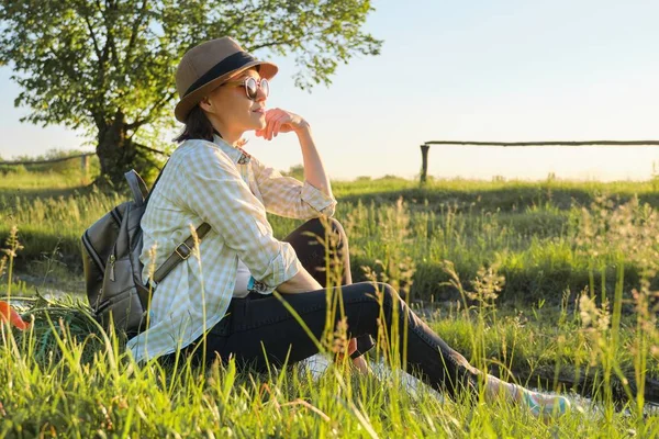 Mujer en sombrero con mochila sentada en la hierba disfrutando de la naturaleza de verano —  Fotos de Stock