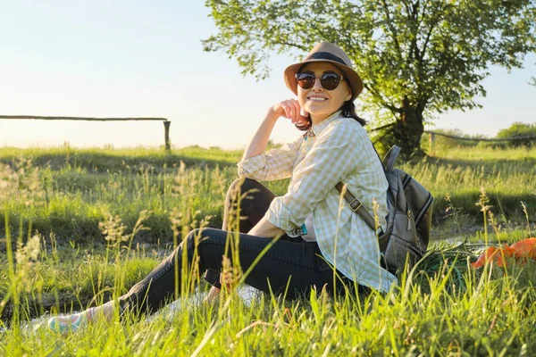 Kvinna i hatt med ryggsäck sitter på gräset njuter av sommaren natur — Stockfoto