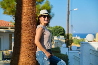 Güneş gözlüklü, olgun, güzel kadın palmiye ağacının yanında duruyor.