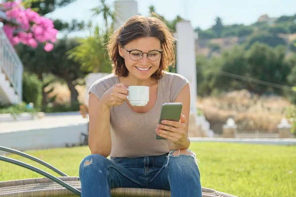 Ώριμη γυναίκα ανάπαυσης υπαίθρια ανάγνωση smartphone με κύπελλο — Φωτογραφία Αρχείου