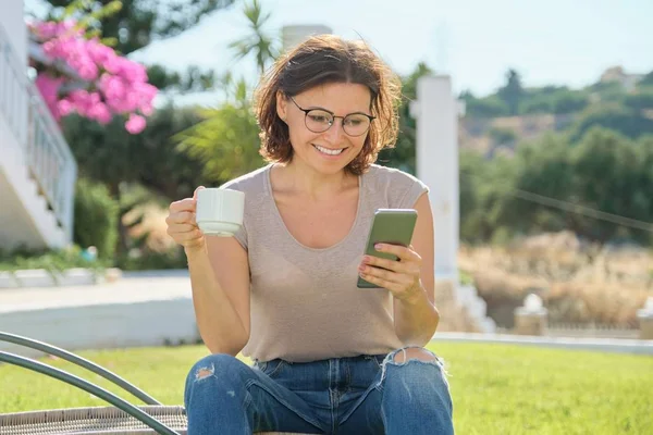 Ώριμη γυναίκα ανάπαυσης υπαίθρια ανάγνωση smartphone με κύπελλο — Φωτογραφία Αρχείου