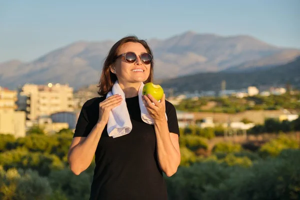 Mature woman in sportswear eating green apple, exercising on nature