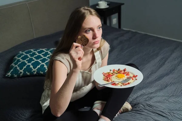 Menina sentada na cama com controle remoto assistindo TV e comendo — Fotografia de Stock