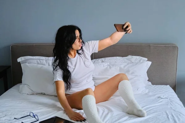 Mulher morena bonita sentada na cama branca levando selfie — Fotografia de Stock