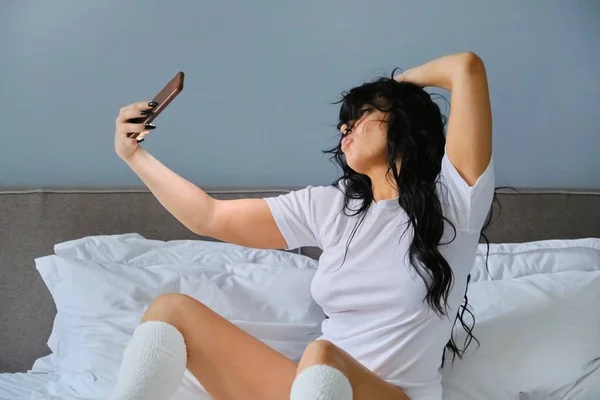 Mulher morena bonita sentada na cama branca levando selfie — Fotografia de Stock