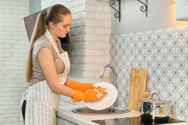 Young woman in apron gloves washing dishes with sponge and detergent