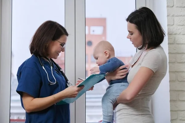 Médico pediatra conversando com a jovem mãe segurando seu bebê em seus braços — Fotografia de Stock