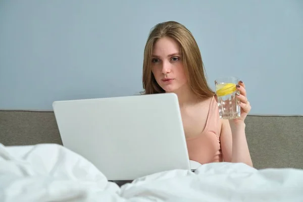 Жінка сидить у ліжку, дивлячись на ноутбук, пити воду з лимоном — стокове фото