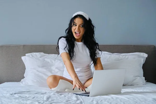 美しいブルネット女性座っているベッドでラップトップコンピュータを使用して — ストック写真