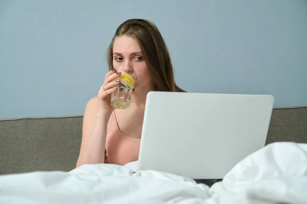 Молода жінка сидить у ліжку з ноутбуком і питною водою з лимоном — стокове фото