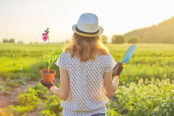 Ung flicka i hatt med handskar går i trädgården med spade och blomma växt i kruka — Stockfoto