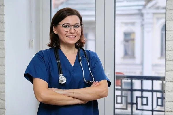 Усміхнена доросла жінка-лікар у синьому однорідному стетоскопі зі складеними руками, впевнена жінка-лікар дивиться на камеру, стоїть біля вікна в клініці — стокове фото