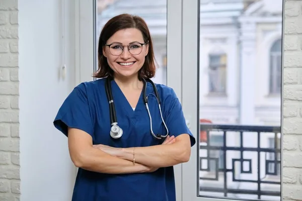 Mavi üniformalı gülümseyen yetişkin kadın doktor kollarını kavuşturmuş, kendinden emin kadın doktor kameraya bakıyor, kliniğin penceresinin yanında duruyor. — Stok fotoğraf