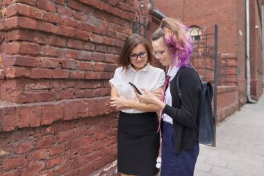 Female student walking and talking with woman teacher, girl showing on smartphone screen