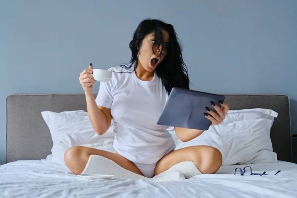 Красива емоційна жінка з відкритим ротом сидить у ліжку за допомогою цифрового планшета — стокове фото