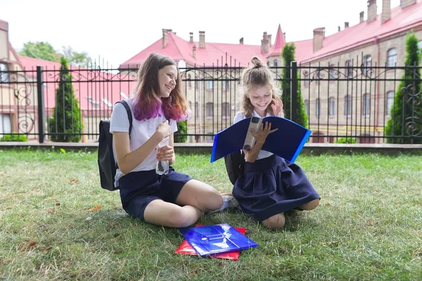 Two schoolgirls sitting on grass with books near school building, girls sisters — 图库照片