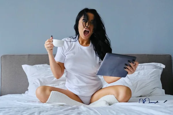 Mooie emotionele vrouw met open mond zittend in bed met behulp van digitale tablet — Stockfoto