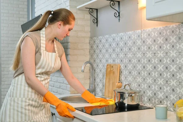 Young woman in apron gloves cleaning washing hob with rag