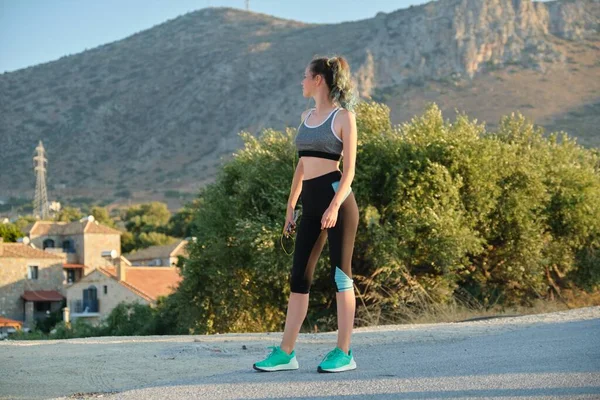 Tiener meisje in sportkleding met smartphone in oortelefoons op de berg weg — Stockfoto