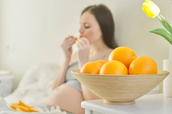 Миска з апельсинами у фокусі, вагітна молода жінка їсть скибочки апельсинів, сидячи в ліжку — стокове фото