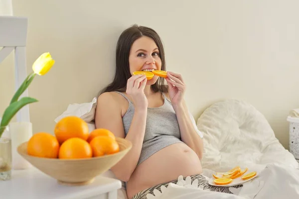 Young pregnant woman eating oranges sitting at home in bed.