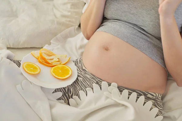 Young pregnant woman eating oranges sitting at home in bed. — Stock fotografie