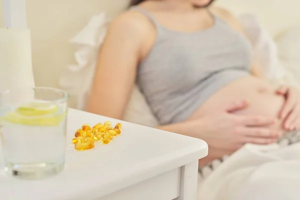 Pregnant female in home interior sitting in bed, vitamin capsules close up — Stock fotografie