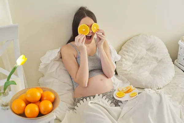 Jovem grávida comendo laranjas sentada em casa na cama . — Fotografia de Stock