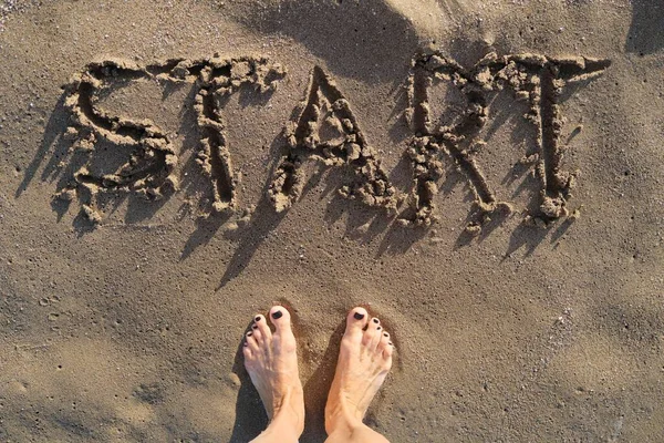 Texto de inicio escrito en arena en la playa del mar y las mujeres pies desnudos — Foto de Stock