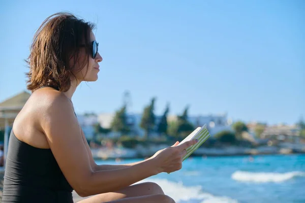 Mujer madura en traje de baño gafas de sol lectura libro sentado en la arena por el mar — Foto de Stock
